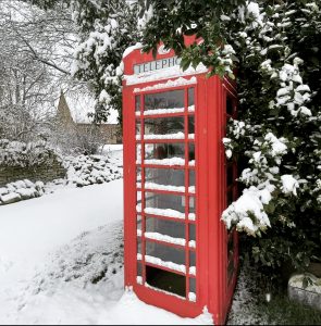 A red telephone box covered in snow with snow covered tracks and cottage behind.