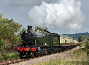 A steam locomotive train, number 280, is puffing steam into the air. The engine is dark green with a black face and red front fender. The carriages are brown and cream. The train is running along tracks in the countryside. The sky is overcast.