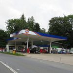 Outside of a Gulf petrol station with some cars at the pumps. The road in front is clear. The sky is overcast. Mature trees are in leaf behind the garage.