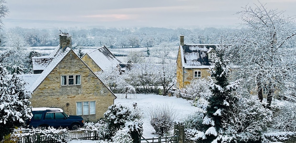 Three Cotswold stone houses can be seen dusted in snow. Beyond them lies snowy open fields and woodland. The pale grey sky has a pink tinge.