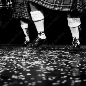 The lower legs of two dancing men can be seen, they are wearing kilts, long white socks, turned over at the top, and black shoes, where the laces are tied around the ankles as well as in the shoes. The floor is black with white speckles.
