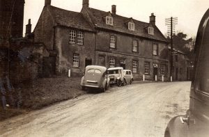 An archive black and white image of the Horse & Groom Pub, Bourton on the Hill. The road leading past seems unsurfaced. Three motor vehicles are parked outside the front, dating from around the late 1930's. Part of a fourth car can be seen to the right, as if the photographer was standing next to it. A tall telecommunication pole is positioned to the right of the pub.