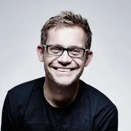 A head and shoulders photograph of a smiling man looking at the camera. He is wearing dark, rectangular framed glasses and a dark round neck jumper. He is in front of a plain light grey background.