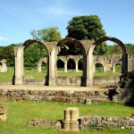 Hailes Abbey ruins are the focal point of the picture, amongst green lawns with trees and blue sky behind.