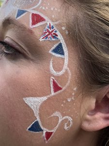 The side of a lady's face has been face painted with curling bunting and white dots. The pennants have been painted in alternate red, white and blue, and one with the union flag. Silver glitter has been added to make it sparkle.