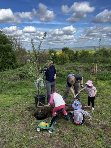 A man and woman help three young children to plant a tree, The lady holds the tree upright while the children shovel earth around its base. They are standing on open ground, over looking a wooded area and views to the countryside beyond. The sky is blue with scudding clouds.