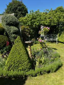 The manicured garden has lawned areas, a pyramid topiary and a bush shaped into a head. A wooden pyramid shaped trellis has pink roses climbing up it. A stone bird bath sits amongst the plants. Mature trees and bushes create a backdrop to the garden.