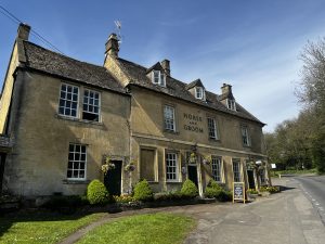 The Horse & Groom Pub stands next to the main road running through Bourton on the Hill. It is built of traditional Cotswold stone. There is a board outside, advertising its real ales and rooms to let. There are small shrubs and grass in front of the pub.