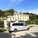 A luxury mini bus stands on the drive outside an impressive Cotswold House, flanked by mature trees and shrubs. The sky is vibrant blue.