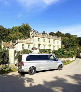 A luxury mini bus stands on the drive outside an impressive Cotswold House, flanked by mature trees and shrubs. The sky is vibrant blue.