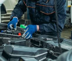 cropped photo showing the torso of a man in dark overalls, wearing blue gloves, leaning over the open engine of a car, touching parts.