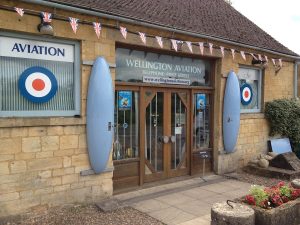 The front door of the Aviation Museum is flanked by aviation parts mounted on the walls. Union Jack bunting hangs above the door. Plant filled stone planters sit at the entrance to the path.