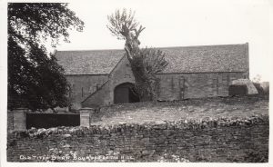 The black and white photo is labelled, 'Old Tithe Barn, Bourton on the Hill', It shows a wide, windowless stone building with a tiled roof. There is a large triangular entrance to the barn with an arched doorway within. Mature trees are on the left and centre of the image. The barn is behind a wall with double wooden gates. The photograph is taken from behind another wall, which appears in the foreground.