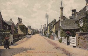 This historic photo is labelled 'Bourton on the Hill - looking up'. It shows the rough road leading up towards the church, with stone cottages lining each side. A woman stands to the left in a blouse and long skirt. The photo would have been taken in the early part of the twentieth century. The original black an white image has had colour added to it.