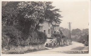 An undated historic photo looking down the main road of Bourton on the Hill. Overhead telecommunication cables can be seen. Cotswold cottages line the left hand side of the picture with steps that lead down from their front doors up the bank straight onto the road.