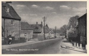 An archive photograph labelled, 'The Hollow, Bourton on the Hill.' The image looks down the main road towards Moreton in Marsh. A girl, with hair in plaits, is running down the left hand pavement. A bench and noticeboard can be seen a little further down the road, There are cottages and houses on either side of the street. A vehicle is driving up the hill, possibly a small bus. The picture is likely to be from the mid twentieth century.