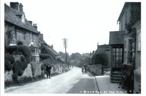 A historic photograph looking down the main road, in Bourton on the Hill. Porch House can be seen to the right. The road is unsurfaced and steps lead down from cottages on the left had bank, straight onto the road. Five people can be seen standing in the road, including a gentleman to the left, two girls pushing a pram. Their clothes suggest the photograph was taken in the early part of the twentieth century.