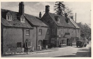 An archive photo labelled, 'Stores & Post Office, Bourton on the Hill'. The photo is taken from a road junction and Cotswold stone properties can be seen to the left. To the middle of the picture sits the village shop. A sign, 'General Stores' hangs above the door with bay windows ether side. The cottages to the left of the shop have white wooden bollards in front of them. A vintage van is parked outside the shop, dating from around the 1930s.