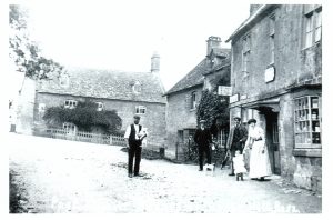 An archive photo of the village Post Office and shop in Bourton on the Hill', looking towards the main road. A sign reading, 'Post Office, Public telephone' hangs from the shop front. A gentlemen in dark trousers and waistcoat, white shirt with rolled up sleeves and a flat cap stands in the unsurfaced road. He is holding something, possibly a dog. Two further men, a young child, a lady and a small dog are standing in front of the shop. Their clothing suggests the picture was taken around 1910. A farmhouse, known as Middle Farm, is seen a the back of the photograph. It has a picket fence in front of it and a shrub growing up the front wall.