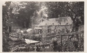 A historic black and white photograph, labelled 'Bourton on the Hill, N'15' shows two joined low Cotswold stone cottages, Rectory Cottage & Slatters Cottage to the right. They have dormer windows in the upper floor. Smoke can be scene coming out of a left hand chimney. Simple gardens are in front of the cottages and mature trees frame the picture, through which the cottages can be seen.