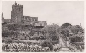 An archive image of St Lawrence Church, labelled,' Rectory Lane, Bourton on the Hill'. Wide, shallow stone steps lead up to an ornate metal pedestrian gate, flanked by cylindrical stone pillars. The initials CEB are just visible at the top of the gate, representing the architect who designed the adjacent gardens and house, Cecil Bateman. Tiered gardens with mature planting lead up to towered church in the the background of the image.