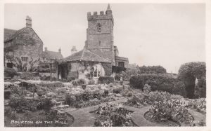 An archive photograph of St Lawrence Church taken from Porch House garden, labelled, 'Bourton on the Hill'. The intricately laid out garden contains low level planting with paved paths and steps leading to different areas. The Cotswold stone Porch House can be seen to the left. There is also a stone built garden studio positioned in front of the church.