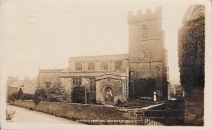 Archive photo of St Lawrence, labelled, 'Church from NW. Bourton. B111' It shows the front of the towered building taken from the road, with a wall in between. Trees and large shrubs are planted outside the church. Notices are pinned to the wooden arch door. The clock on the tower reads 7 minutes past 2.