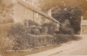 The archive black and white photograph is labelled, 'Bourton on the Hill.' It shows a Cotswold stone cottage to the the left, now known as The Croft. It is fronted with shrubs and flowers. There is an unsurfaced road in front of the cottage. A further property, known as The Bank House, can be seen to the right, with mature trees tower behind it.