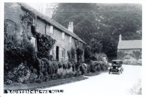 The archive black and white photograph is labelled, 'Bourton on the Hill.' It shows a row of Cotswold stone cottages to the the left, Pilgrims Cottage & The Croft. They are fronted with stone walls with plants growing out of and beneath them. A vintage car is parked on the road, dating from 1920-1930s. A further property, now known as The Bank House, can be seen behind the car. Tall, mature trees tower over it.