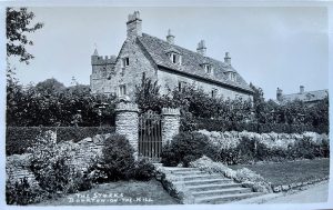 The black and white photograph is of a Cotswold stone property, labelled, 'The Stocks, Bourton on the Hill'. Wide, shallow stone steps lead up to an ornate metal pedestrian gate, flanked by cylindrical stone pillars. Either side are stone walls with grass and shrubs in front. The house is three stories, with three dormer windows in the upper floor. The house has four separate chimney stacks. The Church of St Lawrence is just visible behind the left hand side of the house.