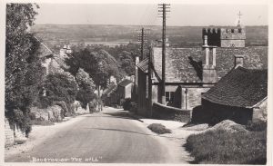 An archive photo labelled 'Bourton on the Hill' looking down the hill towards views over Moreton in Marsh. The village school can be seen on the middle right. Telecommunication poles and cables run down the road. As yet, no pavements have been created on the left, with steps leading down from cottages straight onto the road.