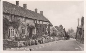The archive photo shows the view looking down the surfaced road of Bourton on the Hill. Pavements can be seen on either side of the road. 'The Bank House' is in the foreground on the left hand side, with a planted front garden and foliage growing up the front of the house,