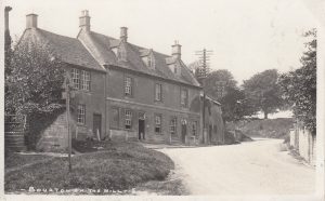 A sepia image of the Horse & Groom Pub in Bourton on the Hill. The road leading past the building is clear and appears to be unsurfaced. It bends round to the left and out of site, There are trees up a bank at the side of the road. Two telecommunication poles are along the road.