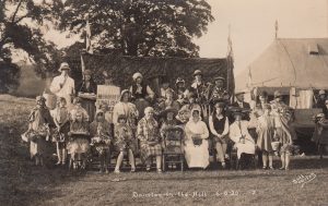 A historic sepia image is of Village residents from who appear to be posing for an amateur dramatics photo, taken by Butt Bourton photographers. Part of a poster can be seen, the words 'Sketch, murdered English' can be seen but the rest is obscured by people. They are in various costumes so it is hard to date the image but probably early 1900s.