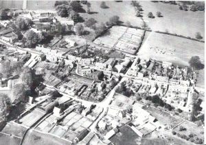 An archive aeriel view of Bourton on the Hill showing the spread of the Cotswold stone houses and cottages on either side of the main road, with farm land behind,