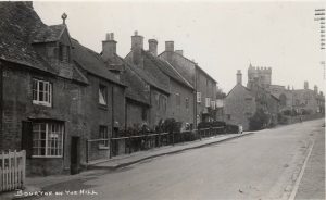 A view of the tarmacked main road running up Bourton on the Hill. Pavements are evident. There are cottages and stone houses on the let side of the photograph, with the church near the top on the left. One of the cottages has a 'Lunches & Teas' sign hanging from it.