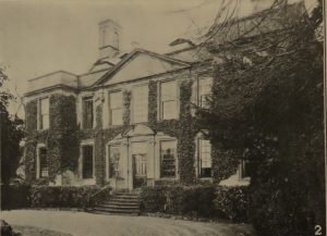 The black and white photograph shows a grand double fronted symmetrical country house. Tall windows grace the two lower of its three floors. Stone steps lead up from a clear driveway to the large front door. Ivy covers most of the front walls but must have been trimmed away around the windows The image is of Bourton House in Bourton on the Hill.