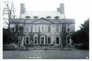 An old photograph, labelled 'Bourton House', which is located in Bourton on the Hill. The black and white image shows a grand three storied house from the rear. It has a symmetrical design with many large windows on the lower ground floors. There is a bare tree to the left, indicating it is winter. There is a low circular stone structure within the lawn, possibly a pond.