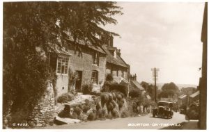 An undated historic photo looking down the main road of Bourton on the Hill with overhead telecommunication cables and an old fashioned car, circa 1920s-30s in the middle ground. Cotswold cottages line the left hand side of the picture.