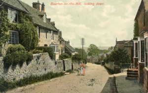 An undated historic picture looking down the unpaved main road of Bourton on the Hill. A woman and child stand in the middle ground. St Lawrence's Church is just visible on the right. Cotswold stone cottages and houses line both sides of the image. The photograph is captioned, 'Bourton on the Hill, looking down'.