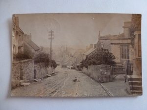 An undated historic picture looking down the currently unpaved main road of Bourton on the Hill. A pony and trap stands in the middle ground. St Lawrence's Church can be seen on the right. Cotswold stone cottages and houses line both sides of the road.
