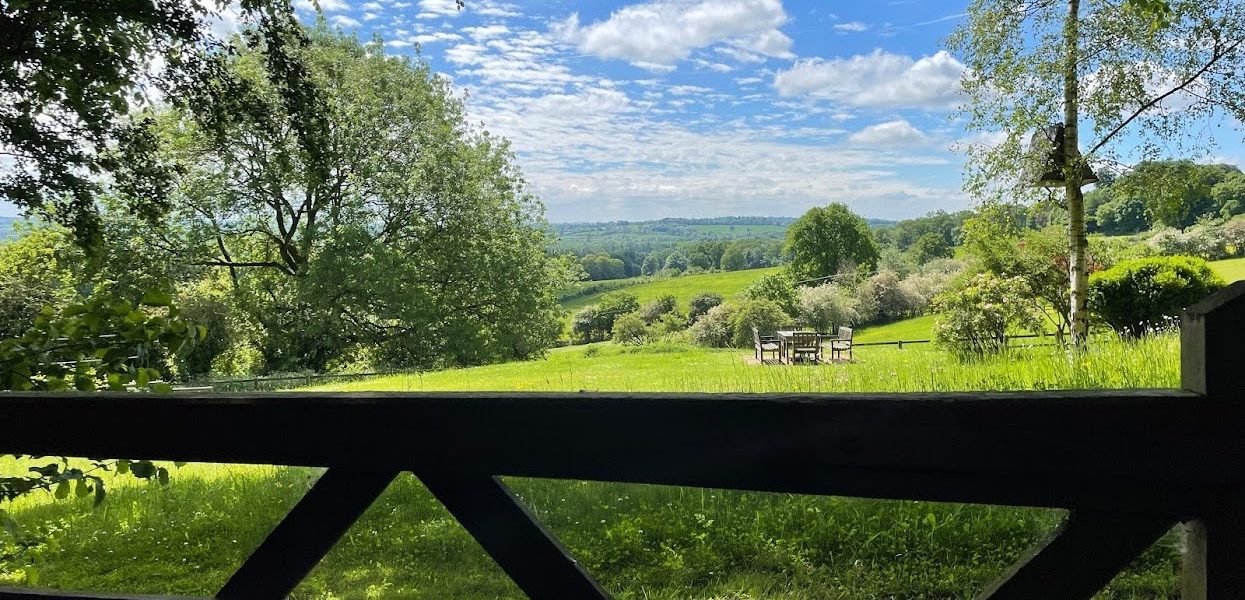 The photo is taken from behind a wooden gate, overlooking a lawned setting with a seating area. Views stretch out across the countryside beyond. The sky is blue with some wispy clouds. Tall trees frame the picture.