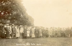 A historic sepia image of a large group of men and women, young and old, dressed in clothes from the early 1900s. They are standing behind a rope in a line. The caption at the bottom reads 'Bourton on the Hill Fete'.