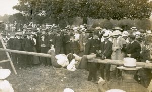 A historic image of a large group of village residents, believed to be taken in 1911. It shows mainly men and boys but a few women are visible. Everyone is wearing hats, the ladies in bonnets and the males in caps or boaters. Everyone is standing in a line watching two males, with their jackets removed, hanging from a long wooden beam in an organised event. A policeman is walking next to the beam.