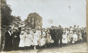 A historic image of at least 60 village residents, men and women, young and old, dressed in clothes from the early 1900s. Most of the young girls are wearing white dresses, the older ladies in black. There is a policeman in the front centre of the group next to a gentleman in a peaked cap. Everyone is wearing hats, the ladies in bonnets and the males in caps. Everyone is standing in a line. The caption at the bottom reads 'Bourton on the Hill Flower Show'.