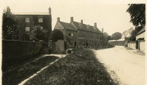 A sepia image of the Horse & Groom Pub in Bourton on the Hill. The road leading past the building is clear and appears to be unsurfaced. Another building, Redesdale House, is to the left of the pub. It is fronted by trees, a wall and steps leading up to a gate at the side. A telecommunication pole is beside the pub.