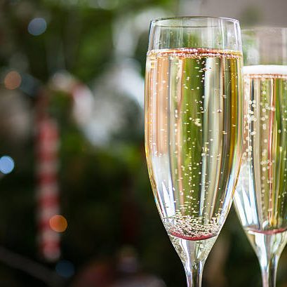 Two tall stemmed flute glasses are filled with sparkling wine. Behind, the image is blurred but shows a Christmas tree with a red and white stripped candy cane hanging from a branch.