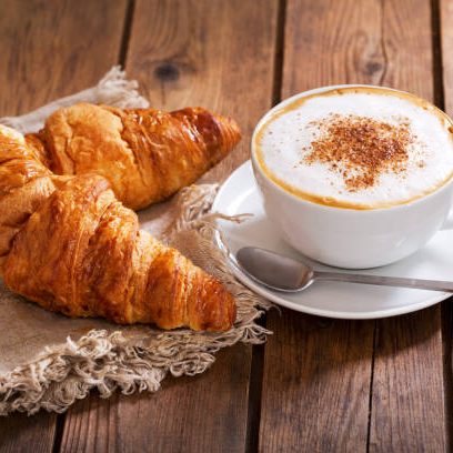 A coffee cup and saucer filled to the brim with frothed milk and a dusting of chocolate powder sit next to a fringed napkin topped with two croissant atop wooden table.