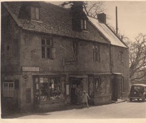 An archive photo of the village Post Office and shop in Bourton on the Hill'. A sign, 'E I Draper, General Stores' hangs above the door with bay windows displaying produce either side. A post office sign is visible above the letter box, set into the wall of the shop. A woman and child can be seen stepping out of the shop. A car is parked to the left, dating from around the 1930s.