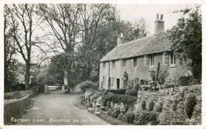 The archive black and white photograph is labelled, 'Rectory Lane, Bourton on the Hill.' It shows a row of Cotswold stone cottages to the right, The Croft & Pilgrims Cottage. They are fronted with stone walls with plants growing out of and beneath them. The road leading past the dwellings has a stone wall to the left. The gates to another property, which cannot be seen, are at the end of the lane, flanked by tall, bare trees.